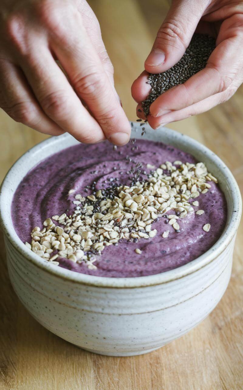 Protein Smoothie Bowl: Your Ultimate Post-Workout Powerhouse 7 Hands adding chia seeds and hemp hearts to a purple smoothie in a white ceramic bowl