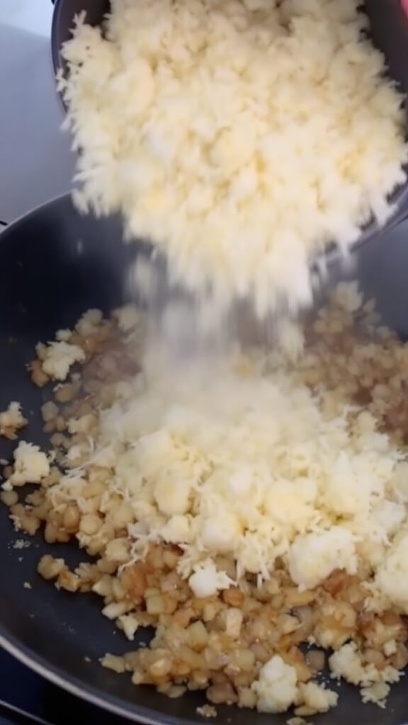 Riced cauliflower being added to skillet with sautéed onions and garlic, steam rising, contrasting colors of white and gold