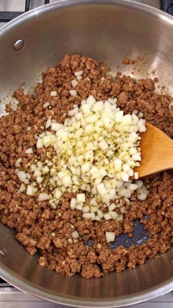 Stuffed Zucchini Boats - A Versatile Low-Carb Dinner 5 Action shot of crumbled italian sausage browning in a large stainless steel skillet over flame, with diced onions and minced garlic being stirred in using a wooden spoon