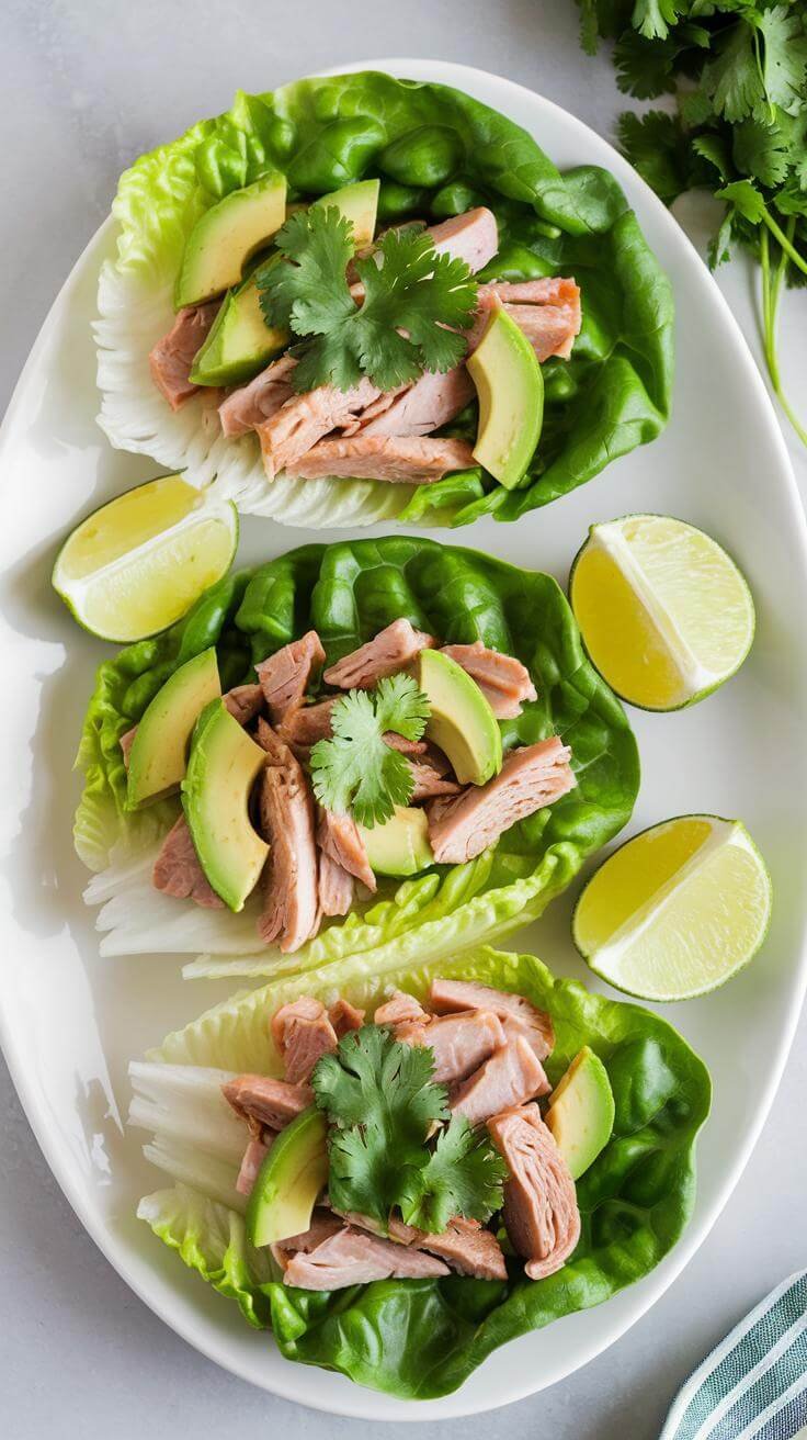 Overhead view of a white plate with three turkey and avocado lettuce wraps, garnished with cilantro and lime wedges.