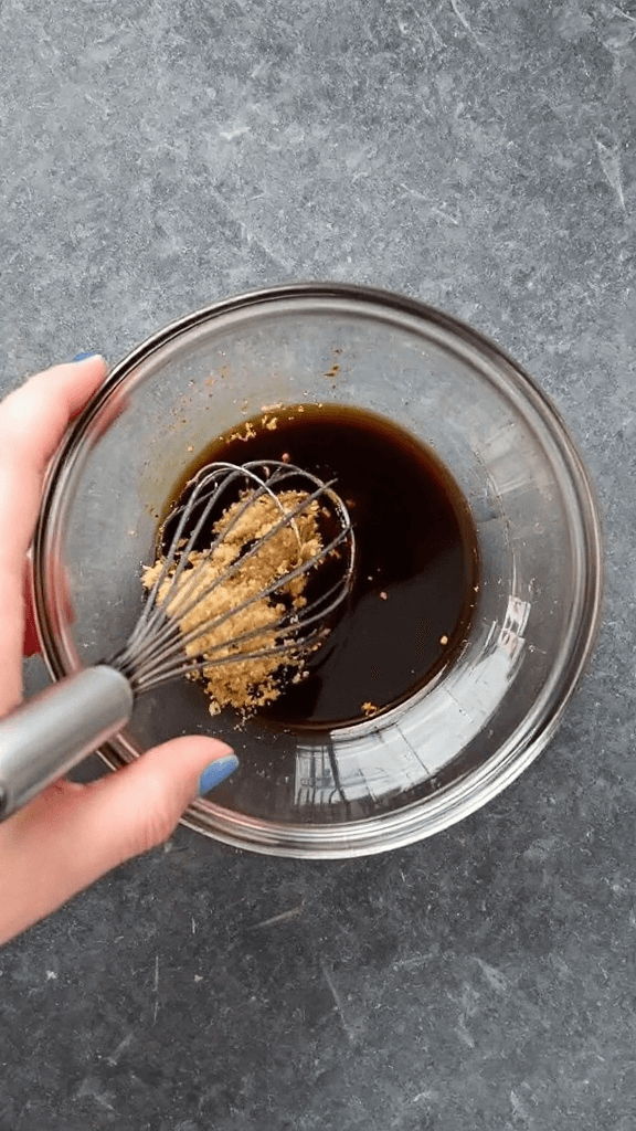 A hand whisking soy sauce, mirin, brown sugar, and sesame oil in a small glass bowl.