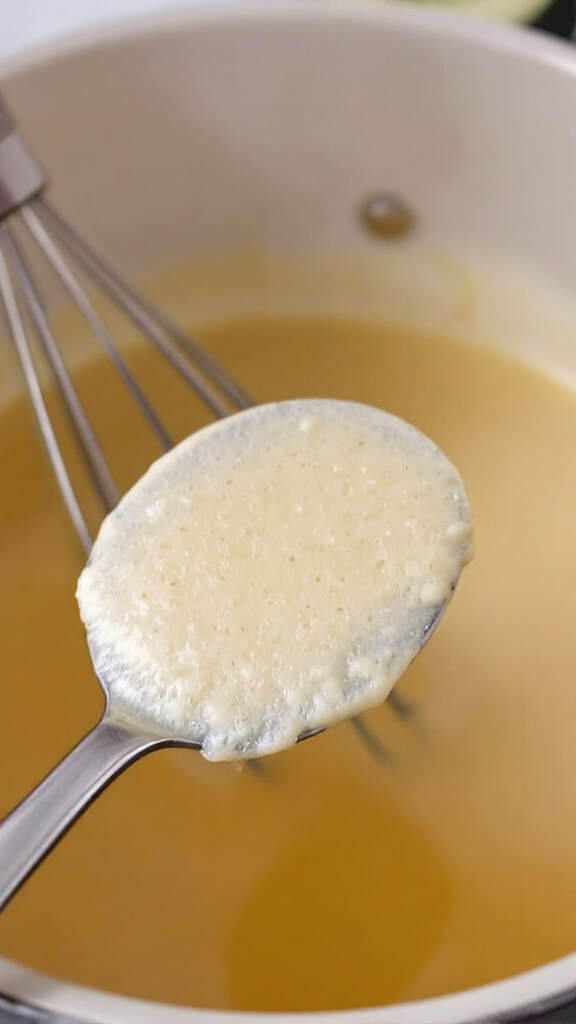 Whisk stirring cornstarch slurry into simmering chicken broth, demonstrating thickening process on a spoon held above the pot
