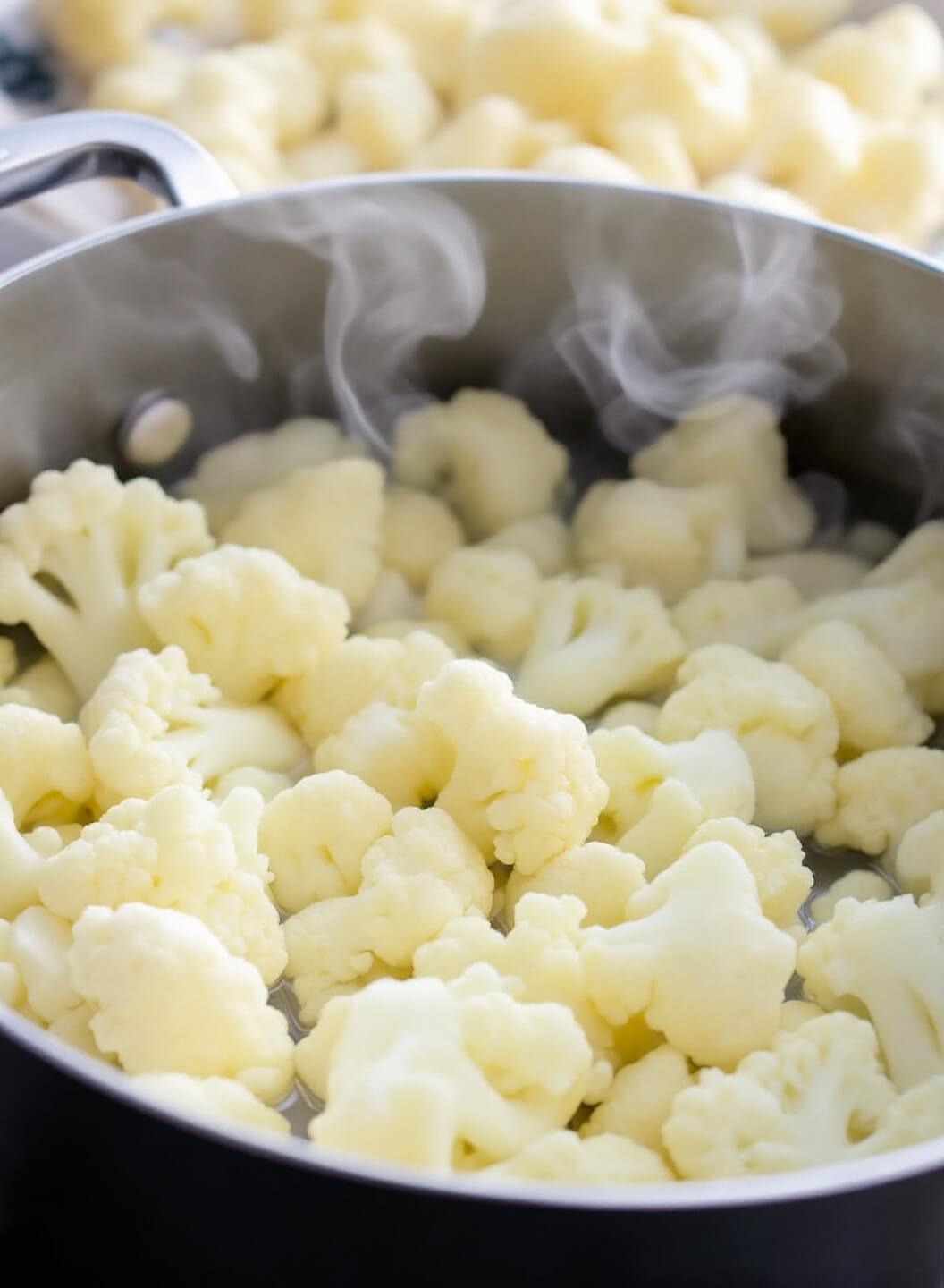 Fresh cauliflower florets boiling in a large pot with steam rising, a colander nearby for draining and the florets appearing bright white and tender