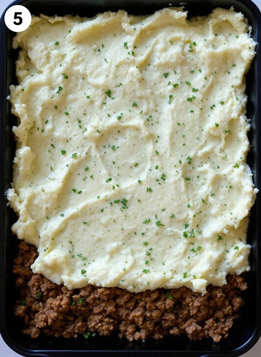Baking dish showing layered meat mixture topped with creamy cauliflower mash, visible herbs included