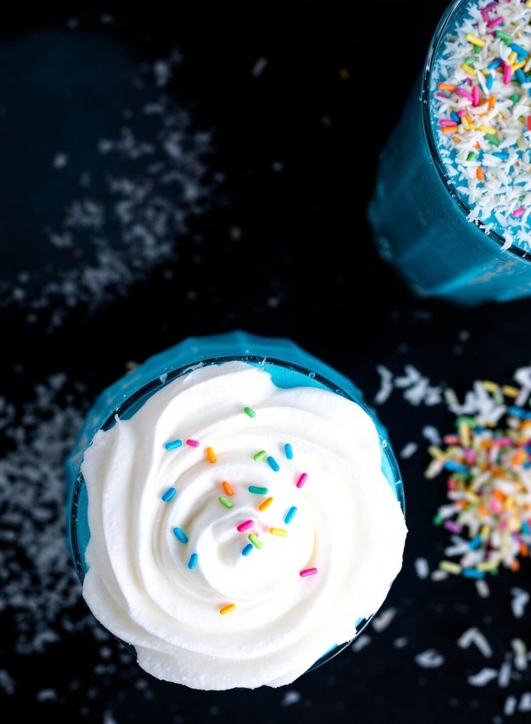 Blue Milkshake Recipe 7 Overhead view of a blue milkshake in a glass, topped with whipped cream, rainbow sprinkles, and coconut shavings.