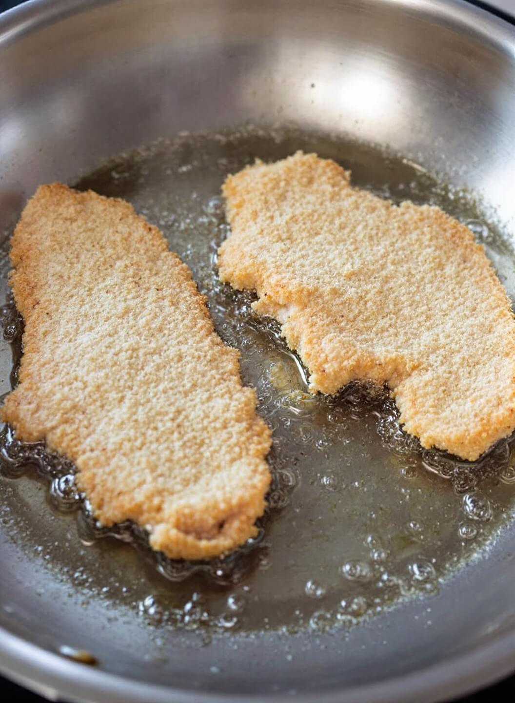 Crispy Buffalo Chicken Breast (3 Ways) 8 Breaded chicken cutlets frying in a large stainless steel skillet, exhibiting bubbling edges and a forming golden-brown crust with steam rising from it