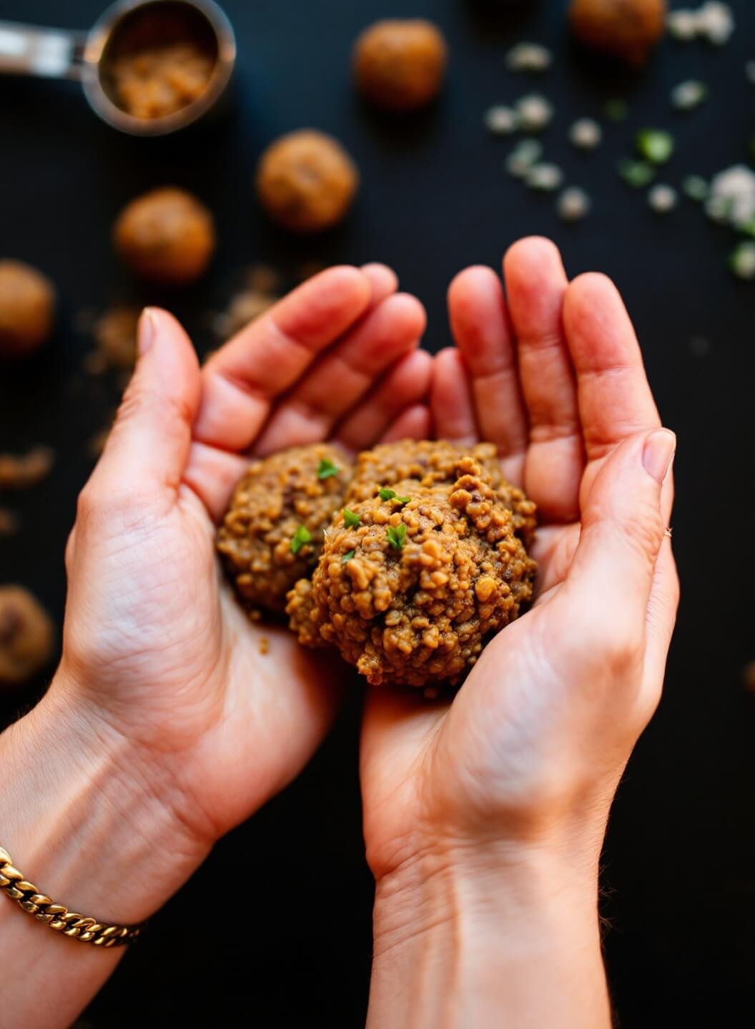 Hands shaping lentil mixture into meatballs on a dark slate surface with cookie scoop and finished meatballs in background
