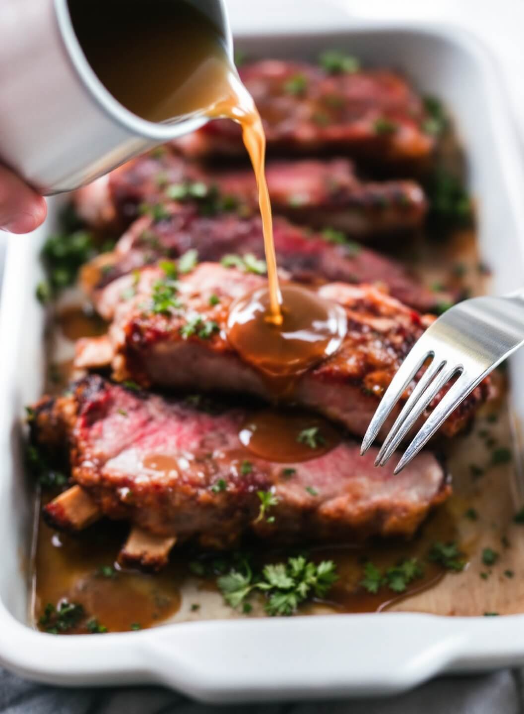 Braising liquid being poured over seared ribs with herbs floating