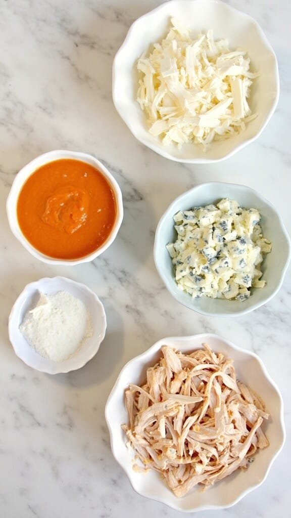Buffalo Chicken Dip With Celery Sticks 3 Ingredients for buffalo chicken dip including cream cheese, buffalo sauce, ranch, blue cheese, and shredded chicken arranged on a marble counter.